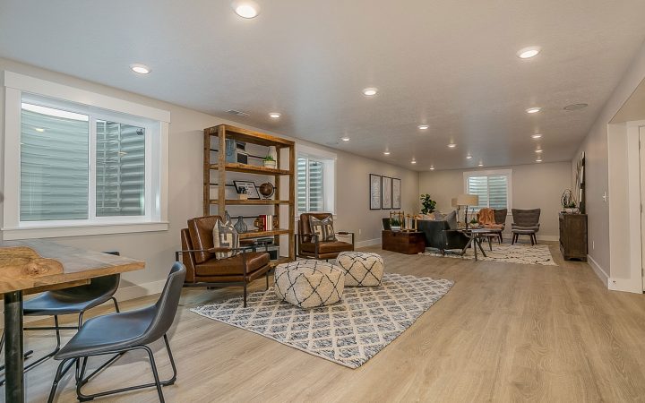 Finished Basement Family Room with Recessed Lighting Fully finished basement recreation room with light wood laminate flooring, multiple seating areas including leather chairs and gray sofas, wooden bookshelf, patterned area rug, recessed ceiling lights throughout, and windows with white shutters