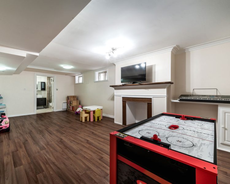 Finished Basement Playroom with Recessed Lighting and Entertainment Area Spacious basement playroom featuring dark wood laminate flooring, red air hockey table, pink toy car, wall-mounted TV above fireplace mantle, electronic keyboard on built-in shelf, children's table and chairs, and recessed ceiling lights throughout