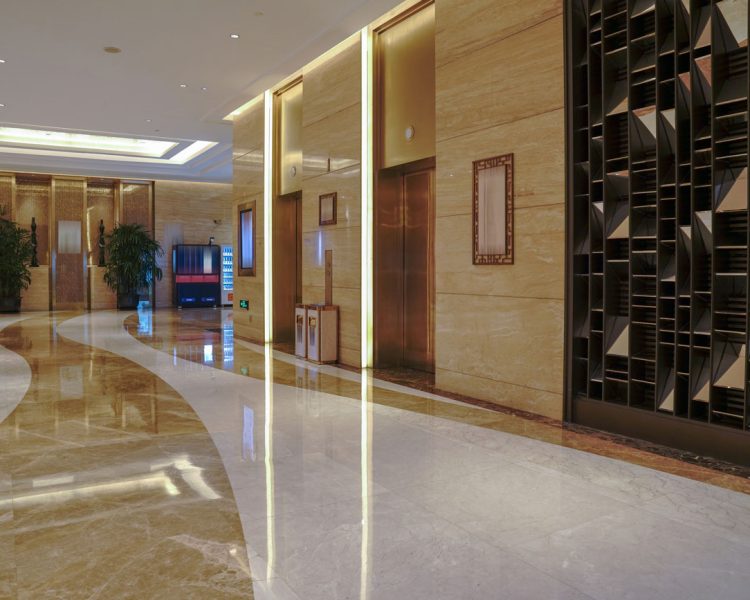 Luxury Hotel Lobby with Elevator Bank Elegant commercial lobby featuring polished marble floors with curved design patterns, golden-toned elevator doors with decorative lighting, beige stone walls, decorative geometric wall panels, and potted plants