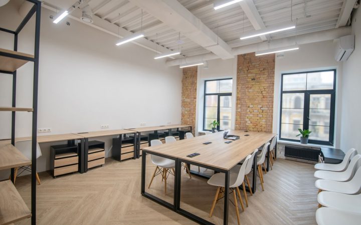 Contemporary Open-Plan Office Space Modern office interior with light wood flooring, long wooden conference tables with white Eames-style chairs, exposed brick accent wall, industrial white ceiling with beam details, large windows, and minimalist black shelving units