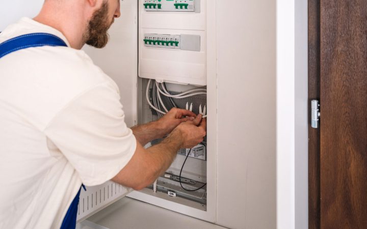 Professional Electrician Working on Circuit Breaker Panel Skilled electrician wearing white shirt and blue suspenders organizing and connecting wiring inside an open electrical breaker panel with green terminal blocks and organized cable management