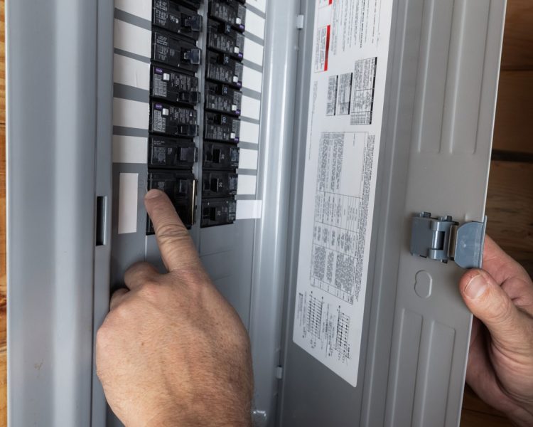 Residential Circuit Breaker Panel Service A close-up view of an electrician's hand flipping a switch on a circuit breaker inside a gray electrical panel box mounted on a wood-paneled wall.