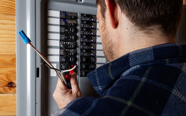 Professional Electrical Wiring and Testing An electrician wearing a plaid shirt using wire strippers to prepare cables while working inside an open circuit breaker panel.