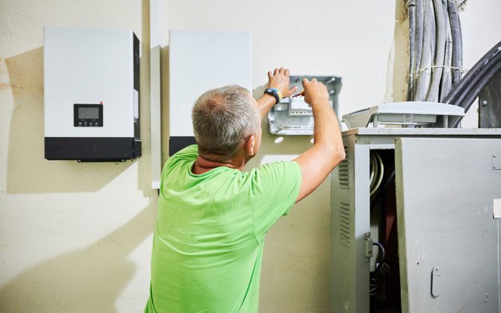 Solar Inverter and Electrical Panel Installation Electrician in bright green shirt installing or servicing wall-mounted solar inverters and electrical distribution equipment with exposed wiring and service panel