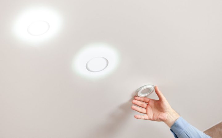 Energy-Efficient LED Recessed Light Upgrade A hand holding a new, unlit LED wafer-style recessed light fixture up to a ceiling where two other identical fixtures are already installed and illuminated, demonstrating a modern lighting upgrade.