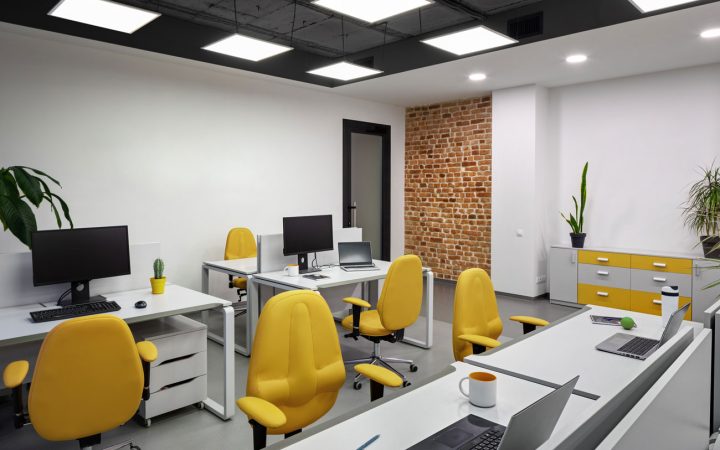 Contemporary Office Space with Modern LED Panel Lighting Bright modern office workspace with white desks, vibrant yellow ergonomic chairs, black exposed ceiling with square LED panel lights, exposed brick accent wall, computer workstations, and indoor plants
