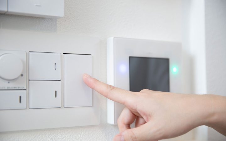 Smart Home Light Switch and Control Panel Installation Close-up of a person's finger pressing a modern white rocker light switch next to a digital smart home control panel with illuminated blue and green indicator lights mounted on a white wall