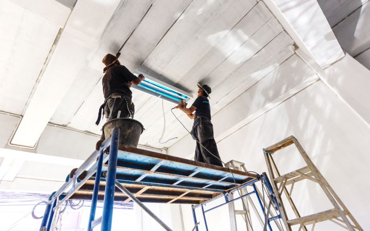 Commercial Fluorescent Lighting Installation Two electricians standing on blue scaffolding installing or servicing fluorescent light fixtures in a high-ceiling commercial building, demonstrating professional commercial electrical contracting services