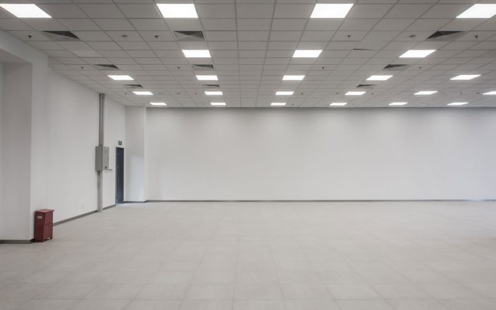 Commercial Office Space LED Lighting Installation Spacious empty commercial interior featuring a suspended grid ceiling with numerous square LED panel lights, white walls, light tile flooring, and a fire extinguisher cabinet.