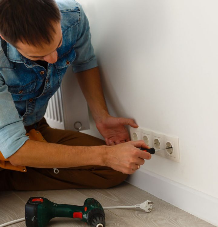 Residential Wall Outlet Installation A handyman in a denim shirt kneels on the floor, using a screwdriver to install a series of white European-style electrical outlets on a clean white wall.