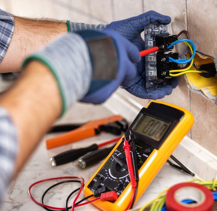 Professional Electrical Outlet Repair and Testing An electrician wearing blue gloves uses a multimeter to test the wiring of an exposed electrical outlet, with various tools like pliers and wire cutters on the marble countertop.
