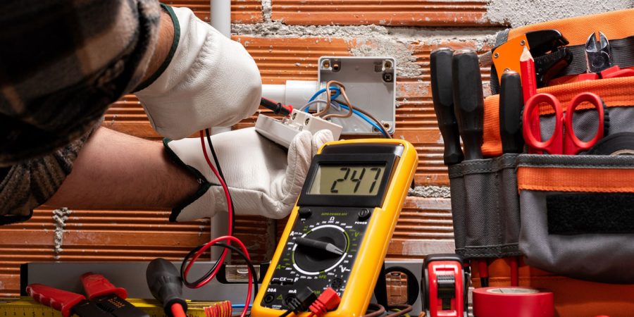 Professional Electrical Testing and Installation Services Electrician wearing white protective gloves using a yellow digital multimeter displaying 24.7 volts while working on electrical wiring in an exposed junction box, surrounded by professional tools including wire strippers, screwdrivers, and an orange tool bag against a brick wall background