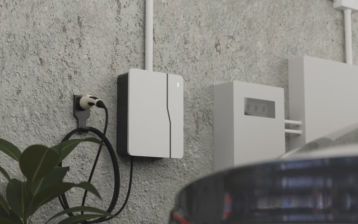 Wall-Mounted Electric Vehicle Charging Station Close-Up Modern white and black EV charger mounted on textured concrete wall with coiled black charging cable and connector, green LED indicator light visible, partial view of electric vehicle in foreground