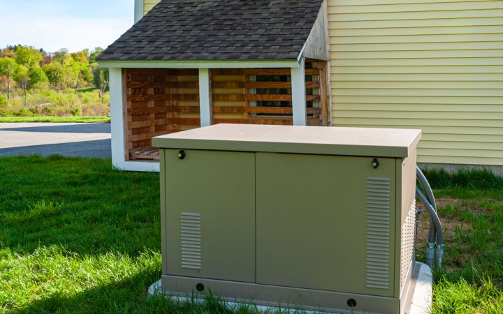 Outdoor Home Backup Generator Setup Tan residential standby generator installed on a concrete pad next to a yellow-sided house, with a wooden shelter structure nearby, set on a green lawn with trees in the background Filename: generator