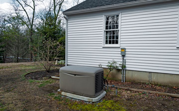 Backup Generator System in a Residential Yard A whole-house generator installed beside a home with white siding, connected to an external utility box, set against a backdrop of trees and a lawn in early spring or late fall.