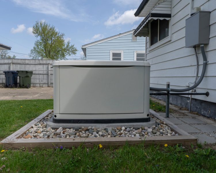 Residential Standby Generator Installation Beige whole-house standby generator installed on a concrete pad surrounded by decorative river rocks in a wooden frame, positioned next to a white vinyl-sided home with electrical conduit connection