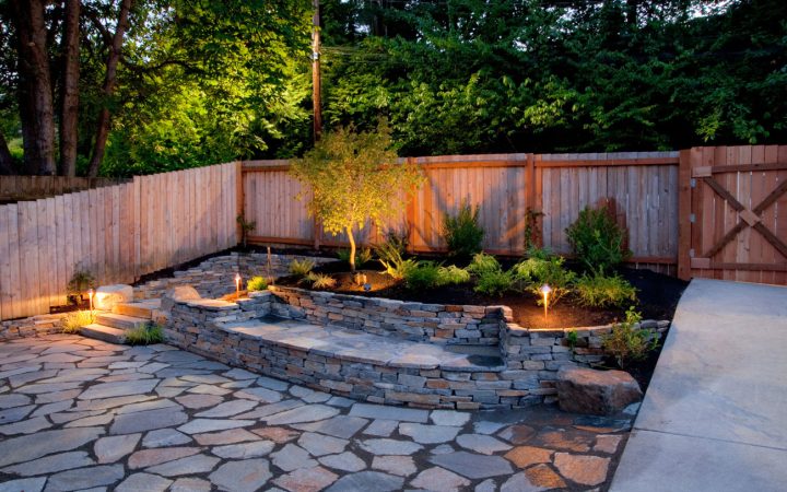 Professionally Lit Backyard Patio and Retaining Wall An elegantly landscaped backyard at twilight featuring a flagstone patio, a tiered stone retaining wall, and strategically placed low-voltage path lights illuminating the plants and walkway.