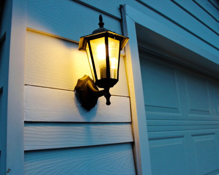 Classic Outdoor Lantern Light Fixture A traditional black metal and glass lantern-style light fixture mounted on white horizontal siding, casting a warm glow at dusk next to a white garage door.